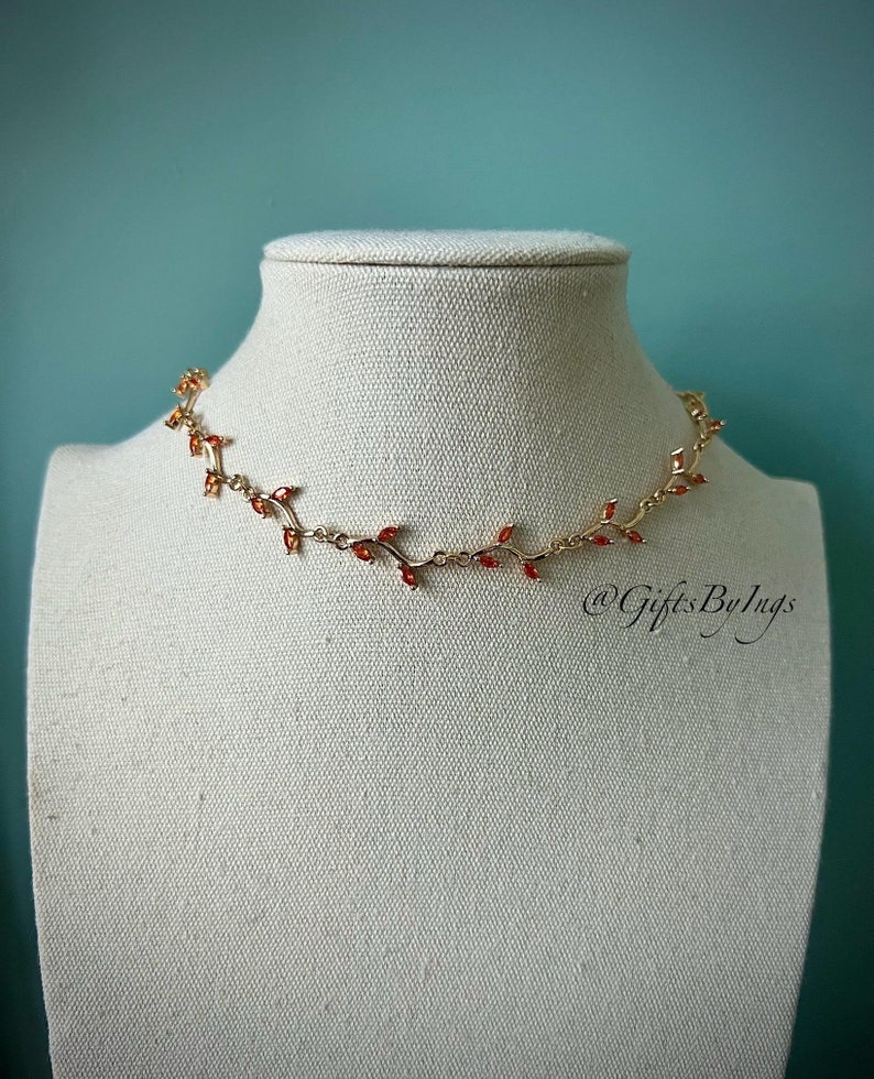 May include: A gold necklace with a delicate vine design featuring small red gemstones. The necklace is displayed on a white mannequin bust.