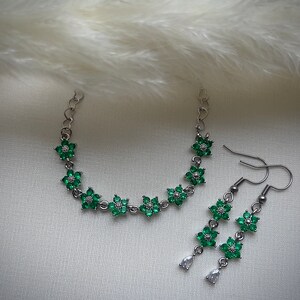 May include: A silver chain bracelet and matching earrings with green flower-shaped charms. The charms are set with clear and green rhinestones.