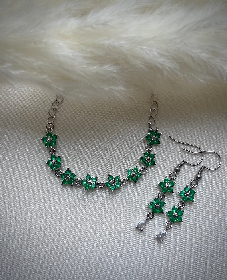 May include: A silver chain bracelet and matching earrings with green flower-shaped charms. The charms are set with clear and green rhinestones.