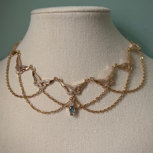 May include: A gold necklace with a delicate, layered chain design. The necklace features a central pendant with a teardrop-shaped blue gemstone and intricate butterfly-shaped accents.