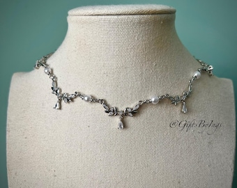 Puede incluir: Un collar plateado con un delicado diseño de hojas y perlas. El collar presenta pequeños cristales transparentes y colgantes en forma de lágrima. El collar se exhibe en un maniquí con un fondo turquesa.