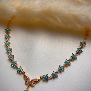 Puede incluir: Un collar de oro con un colgante de mariposa y detalles de flores azules. El colgante tiene una piedra de &oacute;palo en forma de l&aacute;grima.