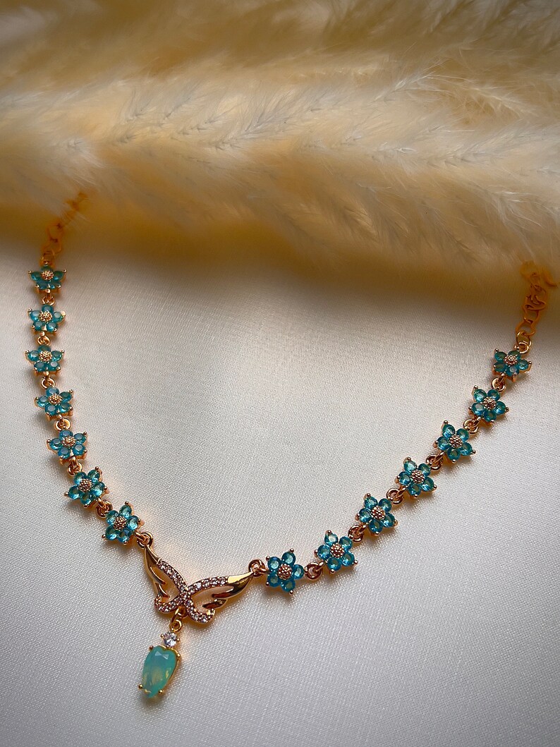 Puede incluir: Un collar de oro con un colgante de mariposa y detalles de flores azules. El colgante tiene una piedra de &oacute;palo en forma de l&aacute;grima.