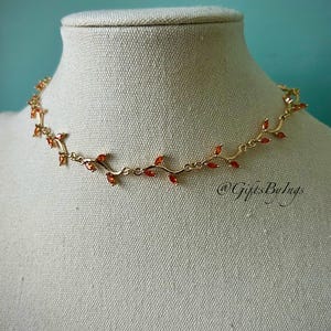 May include: A gold necklace with a vine design and orange-red gemstones. The necklace is on a white mannequin bust.