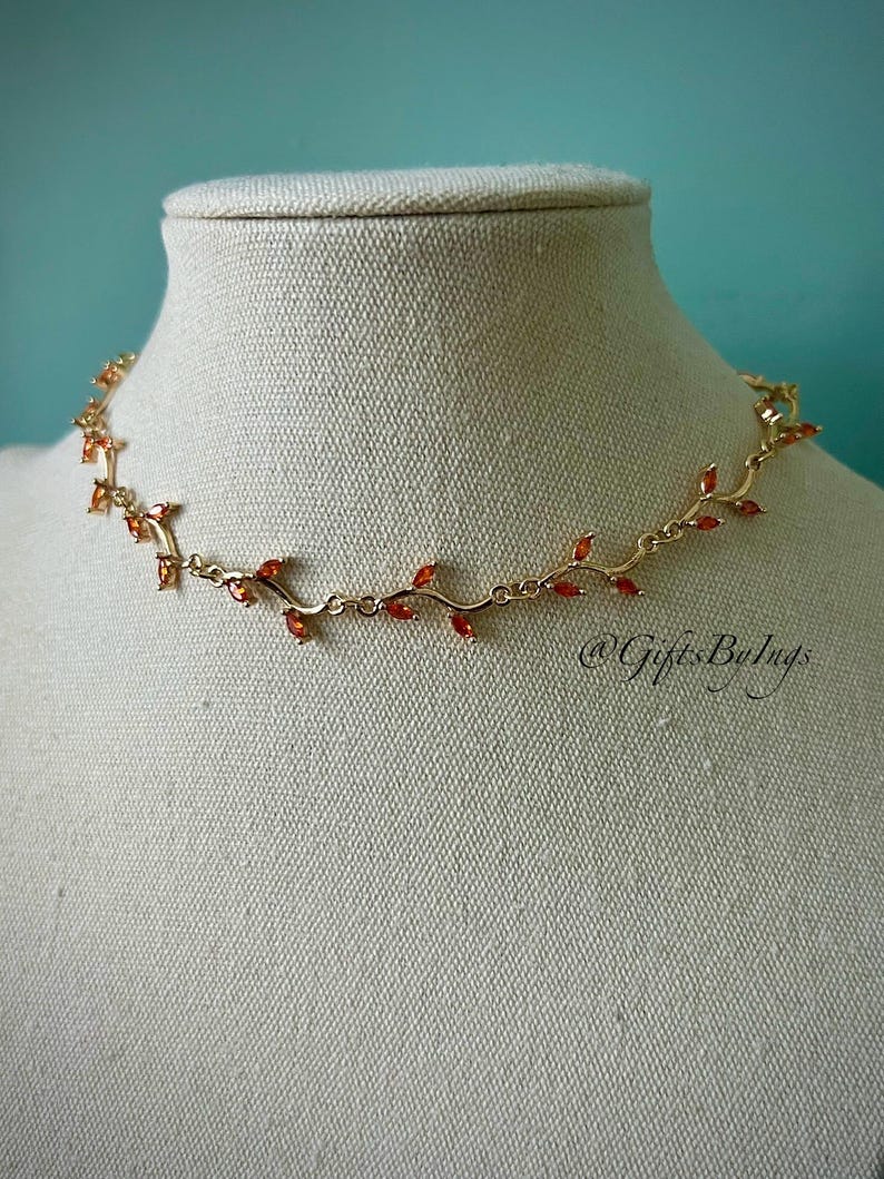 May include: A gold necklace with a vine design and orange-red gemstones. The necklace is on a white mannequin bust.