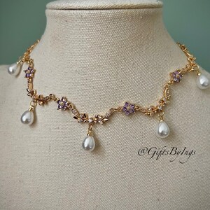 May include: A gold-toned necklace with a floral design, featuring purple flower accents and dangling pear-shaped pearls. The necklace is displayed on a mannequin. The text "@GiftsByIngs" is visible.