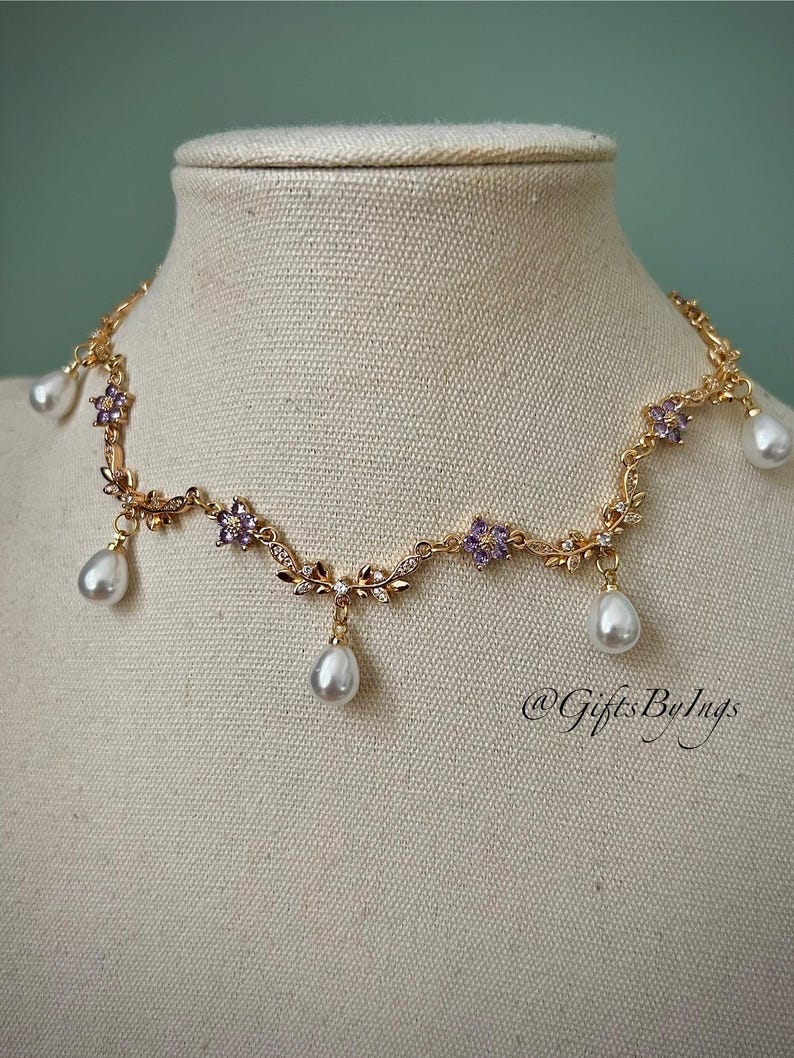May include: A gold-toned necklace with a floral design, featuring purple flower accents and dangling pear-shaped pearls. The necklace is displayed on a mannequin. The text "@GiftsByIngs" is visible.