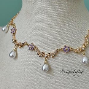 May include: A gold-toned necklace with a floral design, featuring purple gemstone accents and dangling white teardrop-shaped pearls. The necklace is displayed on a mannequin. The text "@GiftsByIngs" is visible.
