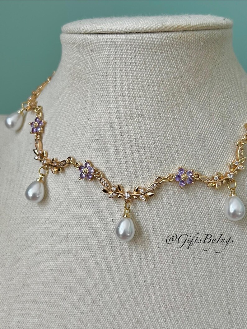 May include: A gold-toned necklace with a floral design, featuring purple gemstone accents and dangling white teardrop-shaped pearls. The necklace is displayed on a mannequin. The text "@GiftsByIngs" is visible.
