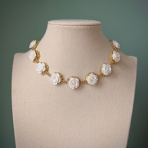 May include: A gold chain necklace with white rose-shaped beads. The necklace is displayed on a white mannequin bust.