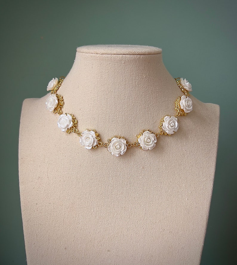 May include: A gold chain necklace with white rose-shaped beads. The necklace is displayed on a white mannequin bust.