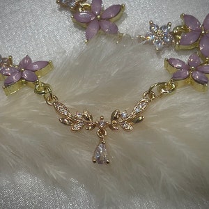 May include: A gold necklace with a delicate floral design and a teardrop pendant. The flowers are pink with clear rhinestones. The necklace is on a white background.