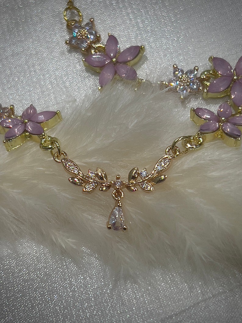 May include: A gold necklace with a delicate floral design and a teardrop pendant. The flowers are pink with clear rhinestones. The necklace is on a white background.