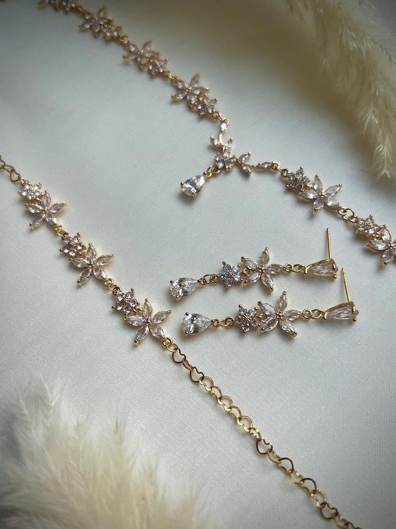 May include: A gold-toned jewelry set consisting of a necklace and a pair of drop earrings. The necklace features a chain with floral motifs set with clear, faceted stones and teardrop-shaped accents. The earrings mirror this floral design.