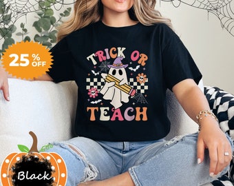 May include: Black t-shirt with the words "Trick or Teach" in retro orange, pink, and white lettering. A ghost wearing a witch hat and holding a pencil is in the center. The shirt has a Halloween theme with bats, flowers, and spiderwebs.