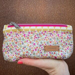 Könnte beinhalten: Eine kleine rosa und gelbe Reißverschlusstasche mit einem weißen Blumenmuster und einem braunen Lederanhänger mit der Aufschrift "jolimop".