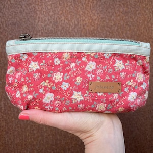 Könnte beinhalten: Eine rechteckige Tasche mit floralem Muster. Die Tasche hat einen roten Hintergrund mit weißen und blauen Blumen. Der obere Rand ist hellblau und weiß gestreifter Stoff. Ein kleines Lederetikett mit dem Wort "jolimop" ist angebracht.