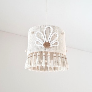 Könnte beinhalten: Eine weiße Makramee-Pendelleuchte. Der Lampenschirm hat ein Blumendesign mit gewebter Seilstruktur, einen zentralen Holzakzent und eine Quastenfranse. Das Licht hängt an einer weißen Deckenleuchte.