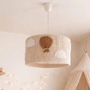 Puede incluir: Un cuarto de bebé en tonos neutros con una cuna blanca, un sillón y una alfombra redonda. La habitación está decorada con peluches, una pantalla de lámpara tejida con un diseño de globo aerostático y un dosel sobre la cuna. El ambiente es cálido.