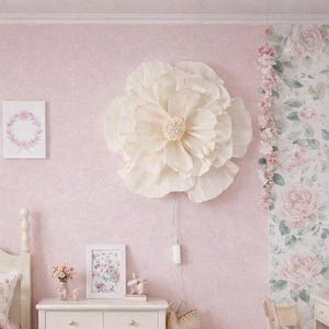 Könnte beinhalten: Ein rosa Schlafzimmer mit weißem Bett, Kommode und Schreibtisch. Die Wände sind rosa gestrichen, die Tapete hat ein Blumenmuster. Eine große, cremefarbene Blumendekoration hängt an der Wand. Ein rosa Teppich und eine Blumengirlande ergänzen die Einrichtung.