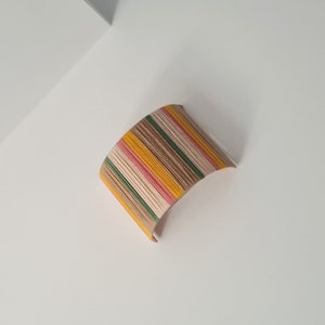 May include: A curved, wooden bangle with a striped pattern in shades of brown, yellow, pink, and green.