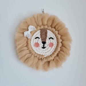 Peut inclure: Une suspension murale tissée beige avec un motif de visage de lion. Le lion a un nez brun, des yeux noirs et des joues roses. Un nœud blanc est placé sur le dessus de la tête du lion.