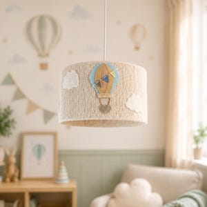 Op de afbeelding: Een beige touwlampenkap met viltapplicaties van een luchtballon en wolken. De luchtballon is blauw, geel en bruin. De lampenkap hangt aan een wit snoer in een kinderkamer.