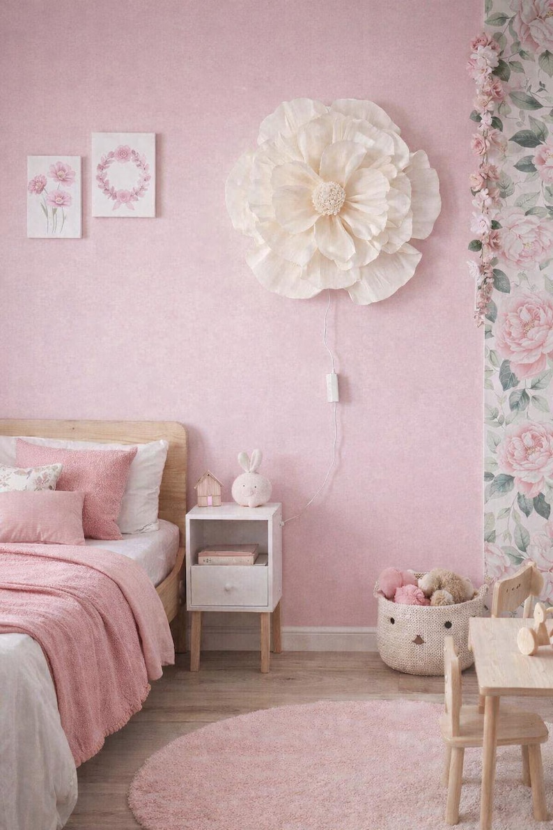 May include: A pink bedroom featuring a large, cream-coloured flower light fixture. The room includes a bed with pink bedding, a white bedside table, and a round pink rug. Two framed pictures hang on the wall.