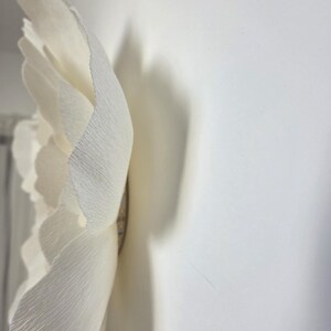 May include: A large, off-white paper flower with textured petals. The flower is mounted on a white wall, casting a shadow. The petals have a ruffled edge, and the centre is a darker shade.