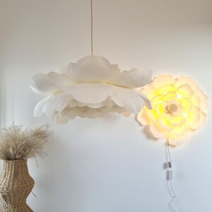 May include: A white, flower-shaped pendant light hangs from the ceiling, with a matching wall-mounted light fixture. The pendant light has multiple layers of petals. A woven vase with dried grass sits below the pendant light.