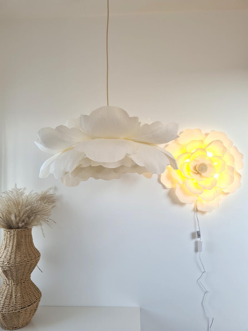 May include: A white, flower-shaped pendant light hangs from the ceiling, with a matching wall-mounted light fixture. The pendant light has multiple layers of petals. A woven vase with dried grass sits below the pendant light.