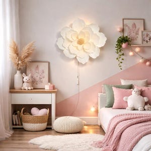 May include: A child's bedroom with a large, white flower-shaped wall light. The room includes a white bedside table, a bed with pink cushions and a blanket, and decorative items such as a bunny plush and framed artwork. The colour scheme is soft, with pink and white accents.