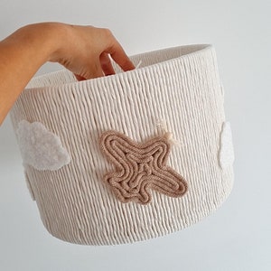 May include: A white woven storage basket with a brown airplane and white clouds. The basket is made of rope and has a textured surface.