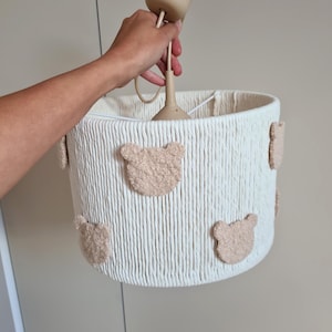 Könnte beinhalten: Cremefarbene Pendelleuchte mit gewebtem Schirm und beige Teddybär-Applikationen. Die Lampe wird von einer Hand gehalten, die das beige Kabel und die zylindrische Oberseite zeigt. Der Schirm ist zylindrisch und aus gewebtem Material.