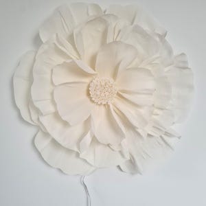 May include: A large, cream-coloured paper flower with multiple layers of petals and a textured centre. The flower is mounted on a white surface, with a thin wire extending from the back. Suitable for wall decor or event decorations.