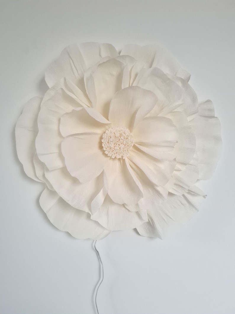 May include: A large, cream-coloured paper flower with multiple layers of petals and a textured centre. The flower is mounted on a white surface, with a thin wire extending from the back. Suitable for wall decor or event decorations.