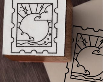 May include: A square wooden rubber stamp featuring a black line art design of a postage stamp with a sun, clouds, and stars. The stamp is shown next to a stamped impression on paper, displaying the same celestial graphic with a scalloped border.