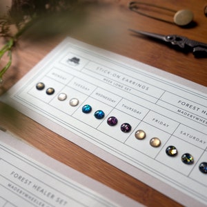 May include: A card displaying a weekly set of stick-on earrings. The card features the days of the week, each with a different colour and style of earring. The earrings are made by "Forest Healer".