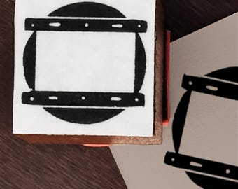 May include: A square wooden rubber stamp featuring a black ink design of a circular shape bisected by two horizontal bars with small rectangular details. The stamp is shown next to a paper impression of the same black ink graphic design.