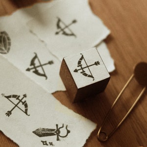 Peut inclure: Un tampon en bois avec une illustration en noir et blanc d'un arc et d'une flèche. Le tampon est posé sur un morceau de papier blanc avec d'autres tampons et une épingle de sûreté.