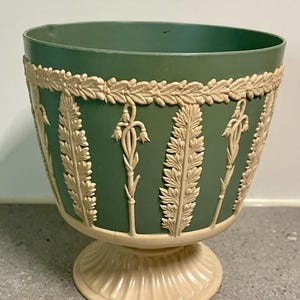 May include: A sage green planter with a cream-coloured base and rim. The exterior features raised cream-coloured relief decorations of vertical fern-like leaves and hanging bell-shaped flowers, with a laurel wreath pattern encircling the top rim.