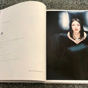 May include: A woman in a black dress with a silver necklace poses for a photo. The background is a dark blue and white gradient.