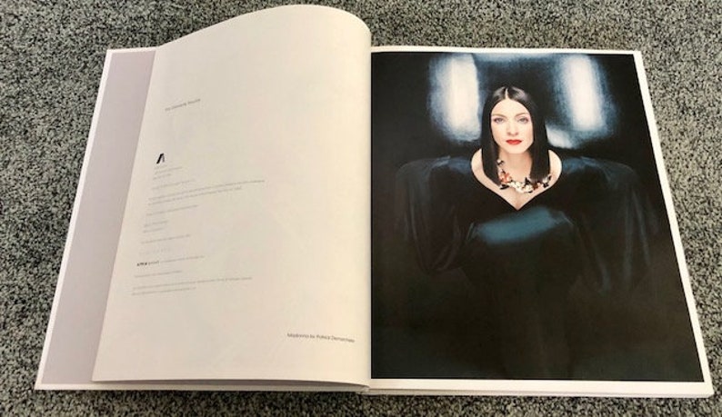 May include: A woman in a black dress with a silver necklace poses for a photo. The background is a dark blue and white gradient.