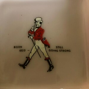 May include: A square, off-white ceramic dish featuring a vintage illustration of a man in a red coat and top hat, walking with a cane. Text reads "BORN 1820 - STILL GOING STRONG."