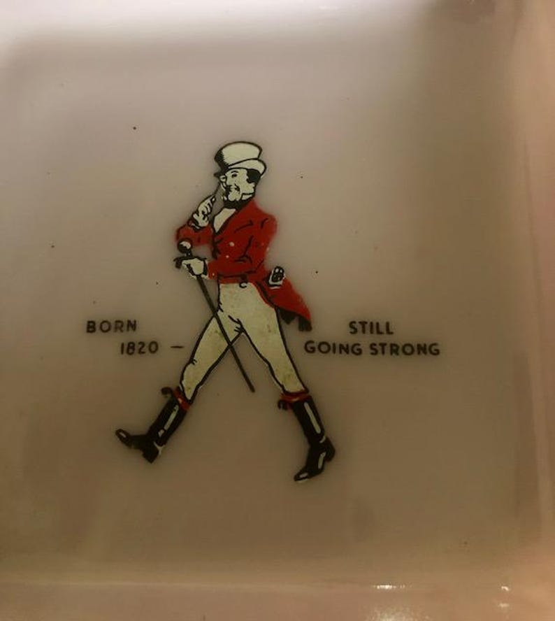 May include: A square, off-white ceramic dish featuring a vintage illustration of a man in a red coat and top hat, walking with a cane. Text reads "BORN 1820 - STILL GOING STRONG."