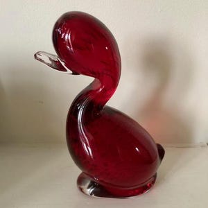 Puede incluir: Una figurita de pato de vidrio rojo. El pato tiene un cuello largo y un cuerpo redondeado.