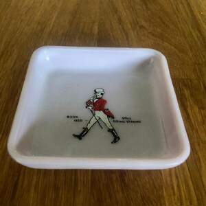 May include: A square, white ceramic tray with a slightly raised rim. The tray features a graphic of a man in a red coat and black boots, with the text "BORN 1820" and "STILL GOING STRONG".