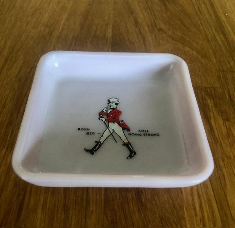 May include: A square, white ceramic tray with a slightly raised rim. The tray features a graphic of a man in a red coat and black boots, with the text "BORN 1820" and "STILL GOING STRONG".