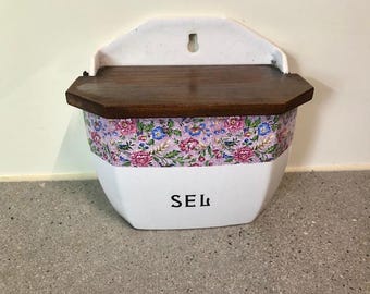 Puede incluir: Salero de cerámica blanca para colgar en la pared con tapa de madera oscura. La caja presenta una banda estampada floral en rosa, azul y verde. La palabra "SEL" está impresa en negro en la parte delantera.