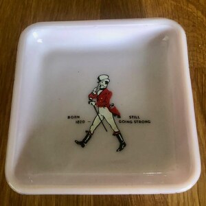 May include: A small, square, pale pink dish with rounded corners. The center features a graphic of a man in a red coat and black boots, with the text "BORN 1820 - STILL GOING STRONG".